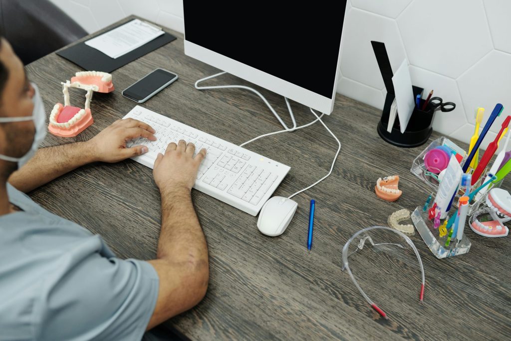 Dentist at desk typing on computer with dental models and tools around.